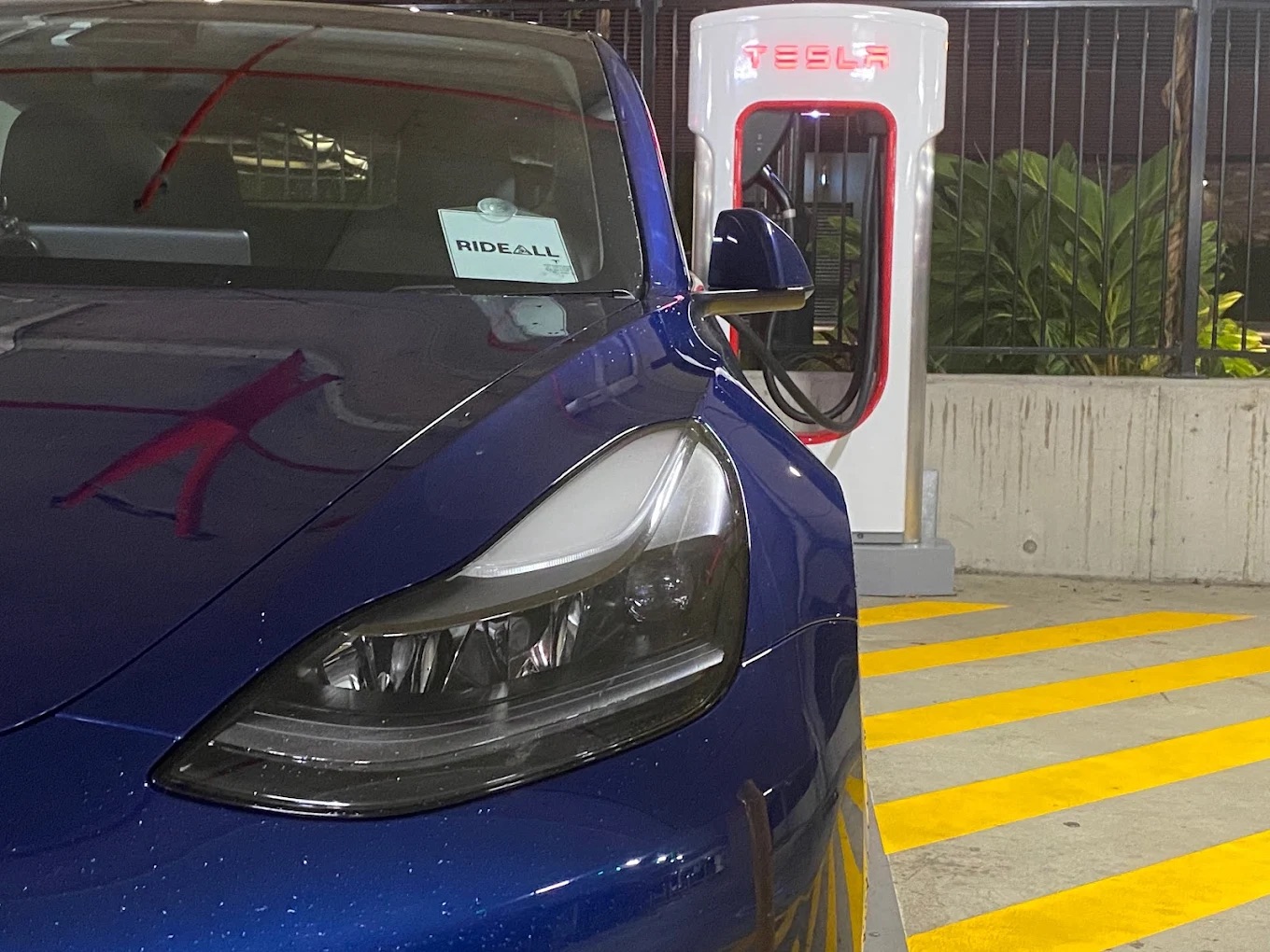 RideAll's blue Tesla parked beside a Tesla charger, with a RideAll sign on the dashboard